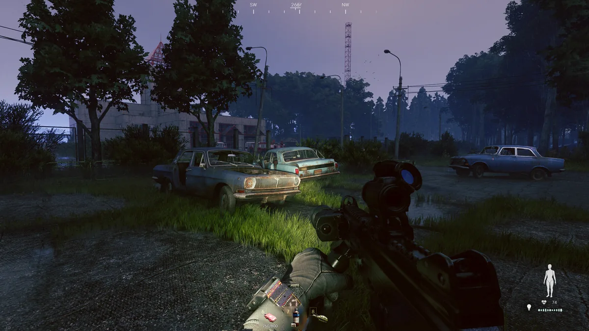 A player character in Soviet-military gear moves through dusk grass toward abandoned sedans near a fenced research compound, rifle scope raised