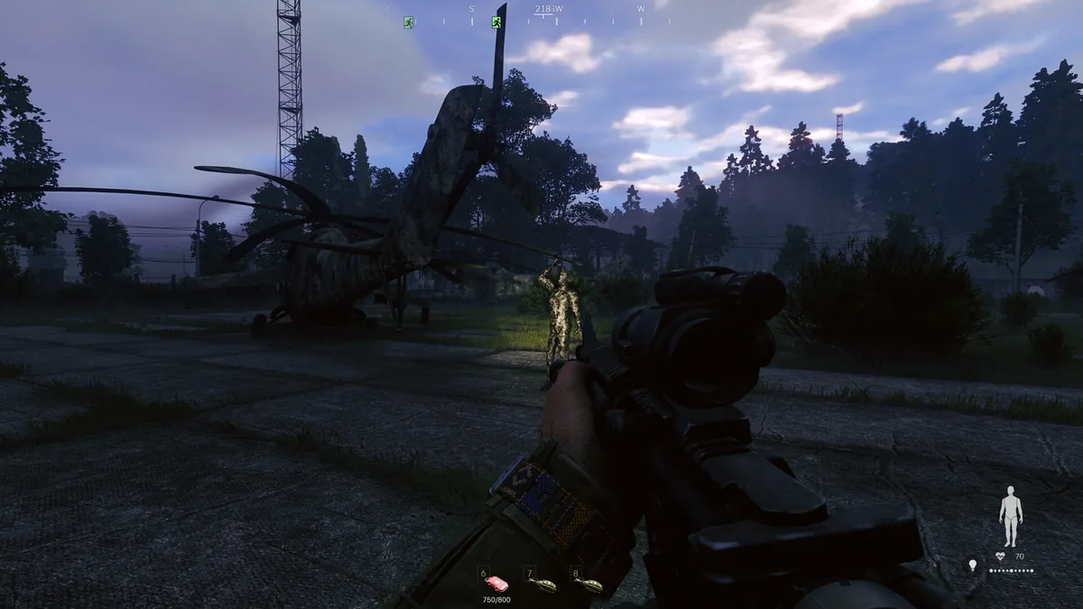 A player aims a scoped rifle at another camouflaged operative near a crashed helicopter on an airfield at dusk
