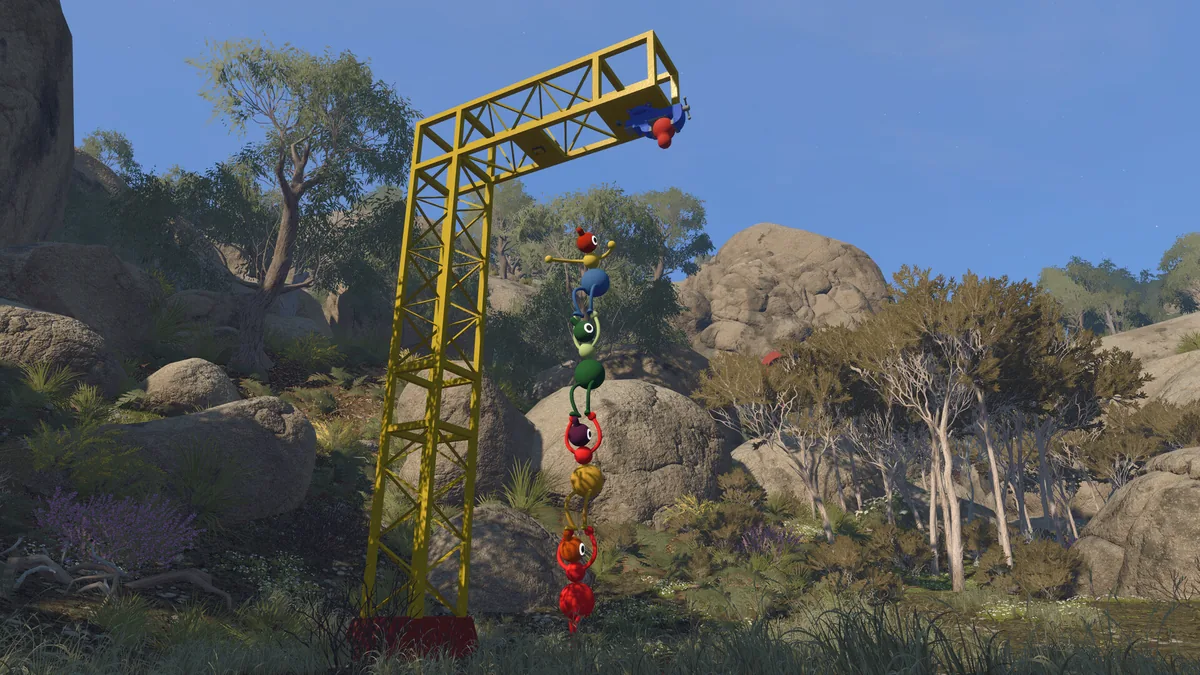 A tall stack of Big Walk characters forming a chain up toward a yellow crane arm against a clear blue sky and rocky hillside, with the stack's reflection visible below