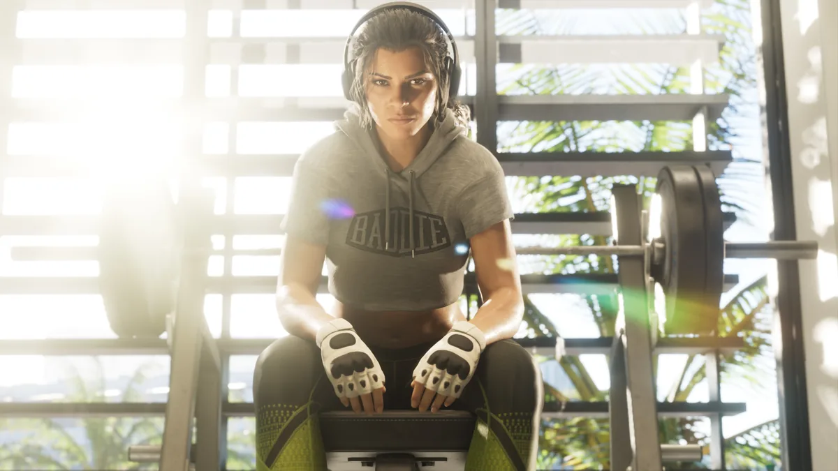 Lucia Caminos wearing a gray Baddie hoodie and workout gloves at a home gym bench, palm trees visible through the blinds behind her