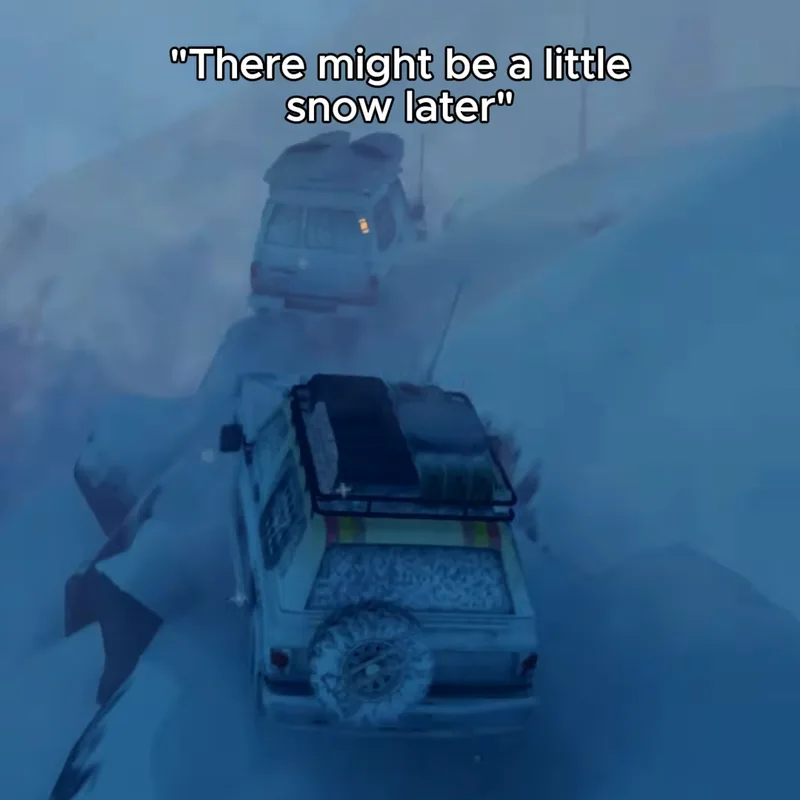 A white 4x4 with a roof rack and spare wheel crawls along a snowy mountain ledge, almost hidden by blue snowfall