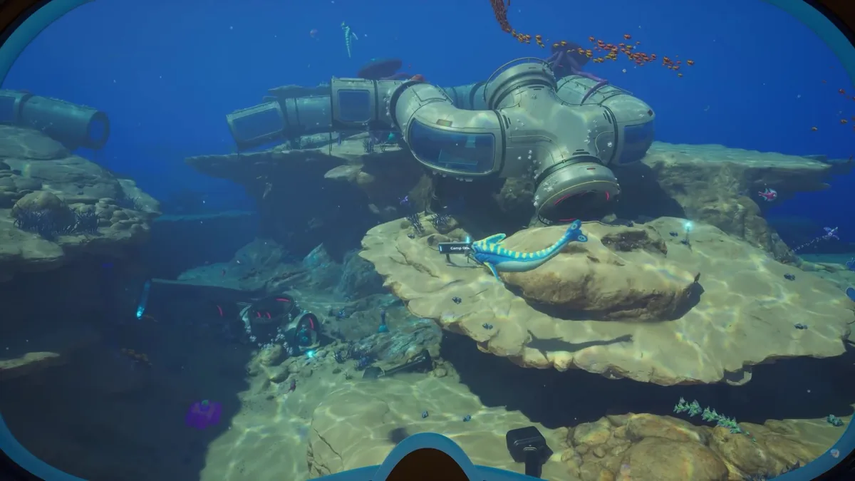 Diver approaching salvaged wreckage and machinery resting on a rocky ledge, a small striped fish nearby and a partial location label visible