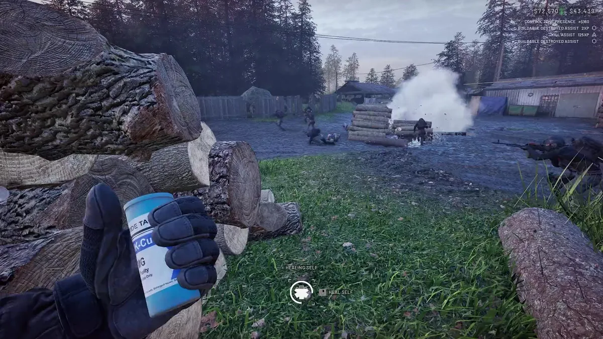 First-person view holding a can of medication with a HEALING SELF prompt, log piles and a barbed-wire-topped fence nearby, smoke rising from a firefight in the background