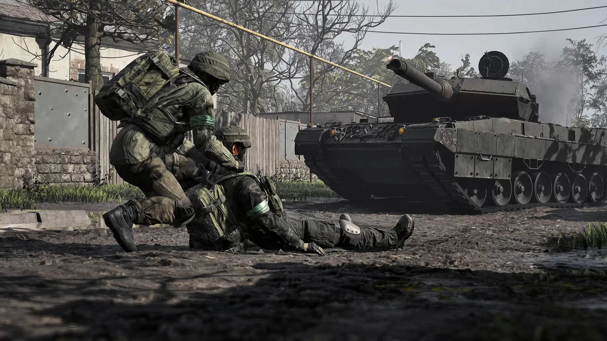 Two soldiers help a downed squadmate to their feet on a muddy street as an armored tank fires in the background near a residential fence line