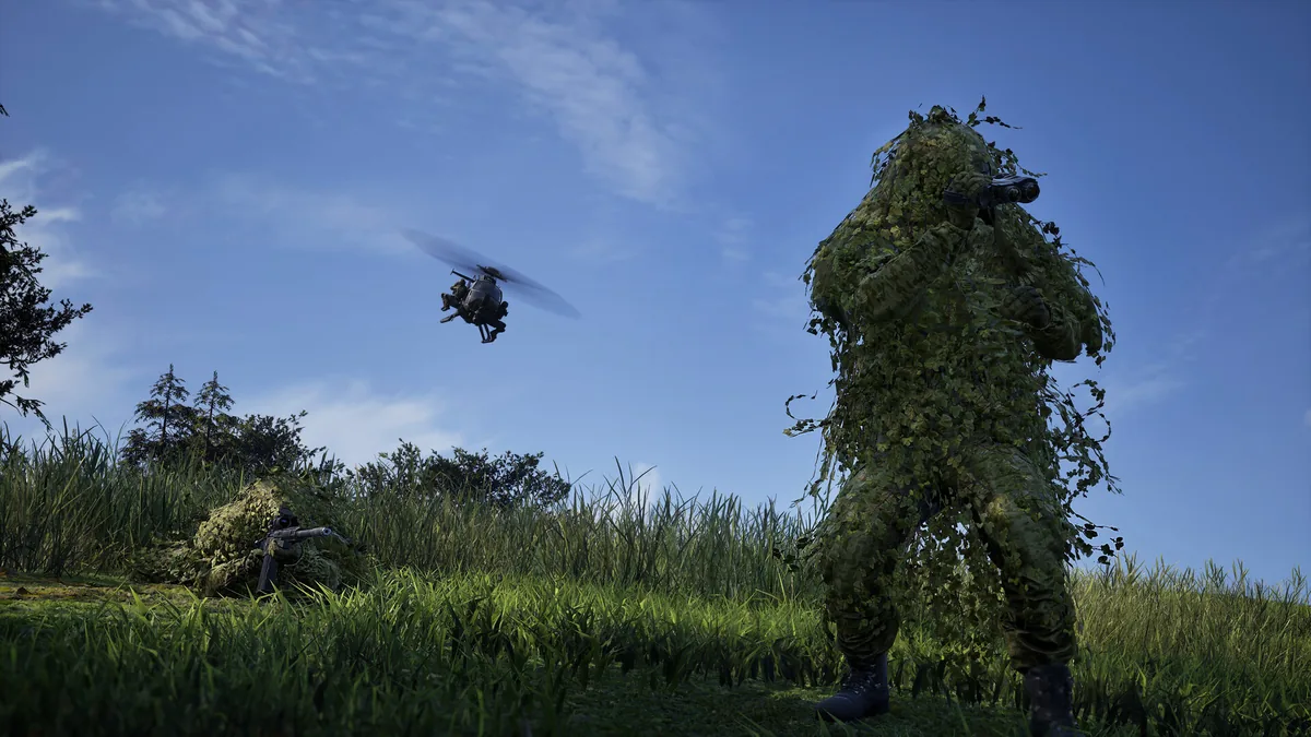 A ghillie-suited sniper aims binoculars skyward while a second ghillie-suited teammate lies prone with a rifle, a scout helicopter passing overhead against a clear blue sky